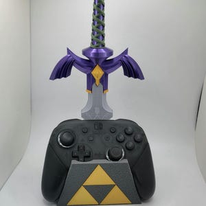 May include: A purple and gold Master Sword video game controller stand with a Nintendo Switch logo. The stand is designed to resemble the Master Sword from the Legend of Zelda video game series.
