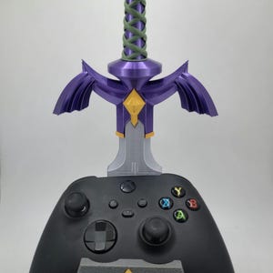 May include: A black video game controller with a purple and silver Master Sword stand. The stand features a triangular base with a gold Triforce symbol.