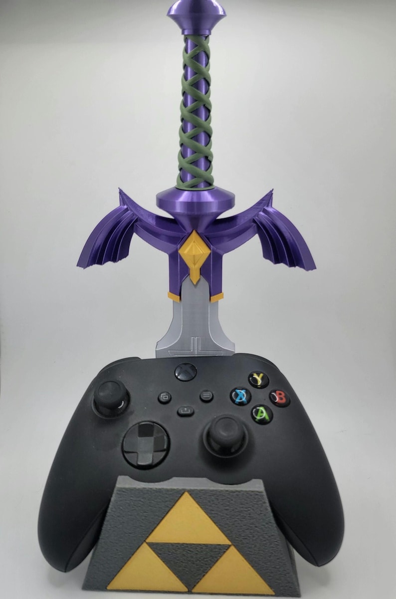 May include: A black video game controller with a purple and silver Master Sword stand. The stand features a triangular base with a gold Triforce symbol.