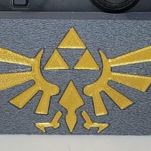 May include: A gray and gold video game controller stand with a Triforce design. The stand is made of plastic and has a textured surface.