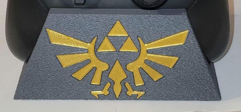 May include: A gray and gold video game controller stand with a Triforce design. The stand is made of plastic and has a textured surface.