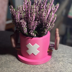 Puede incluir: Una maceta rosa con un diseño de cruz blanca, llena de plantas de flores moradas. La maceta tiene asas marrones y una base a juego, colocada sobre una superficie gris. El diseño general recuerda a un personaje de dibujos animados.