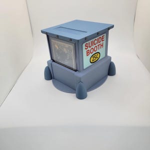 Op de afbeelding: Een lichtblauwe, doosvormige display met een kaart zichtbaar aan de binnenkant. De behuizing heeft de woorden "SUICIDE BOOTH" en "25&cent;" op de zijkant gedrukt. De behuizing heeft een platte bovenkant en vier afgeronde voetjes.