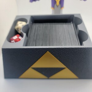 May include: A gray card game holder with a gold triangular design. The holder has a compartment for cards and a separate compartment for dice.