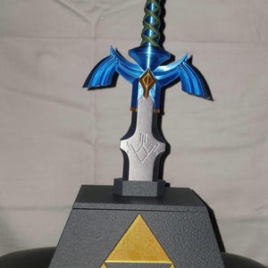 May include: A blue and silver Master Sword replica on a black pedestal with a gold Triforce symbol. The sword has a blue handle with a green braided wrap.
