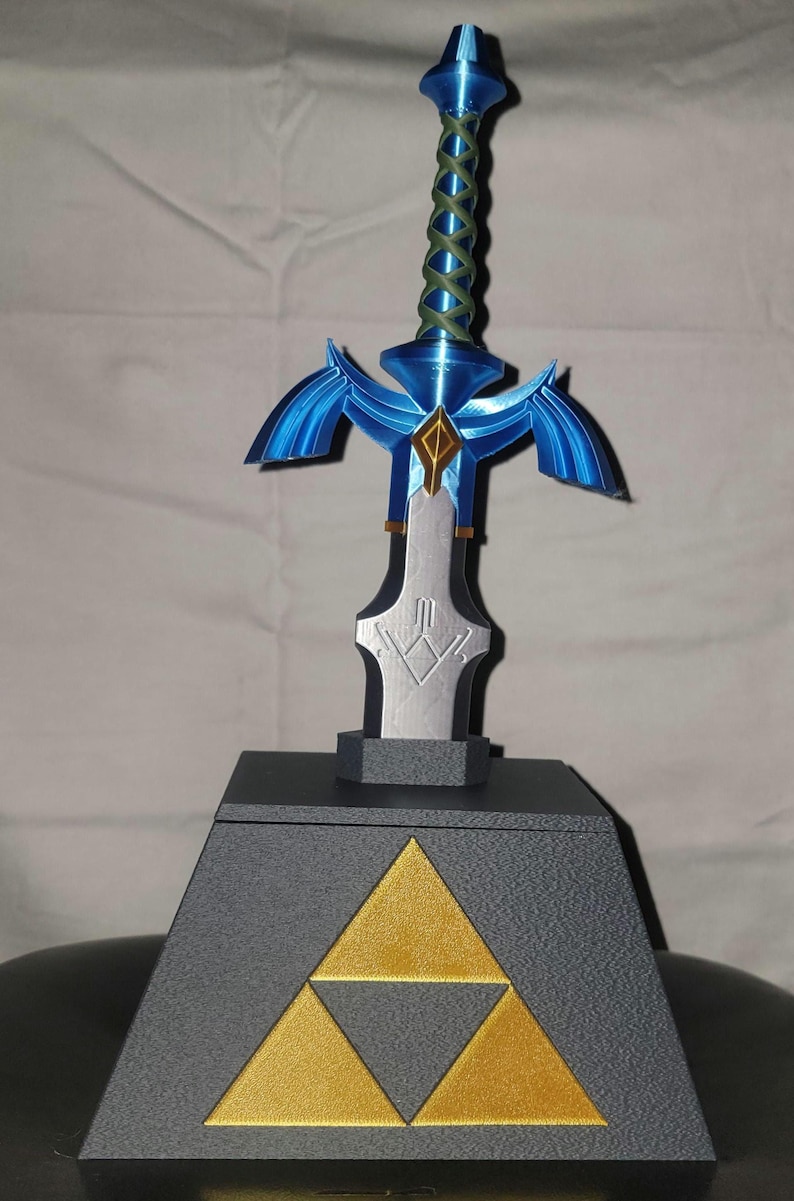 May include: A blue and silver Master Sword replica on a black pedestal with a gold Triforce symbol. The sword has a blue handle with a green braided wrap.