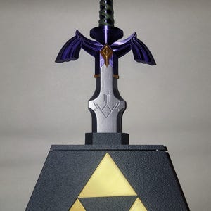 May include: A purple and silver Master Sword replica with a green rope handle, standing on a black base with a gold Triforce symbol.