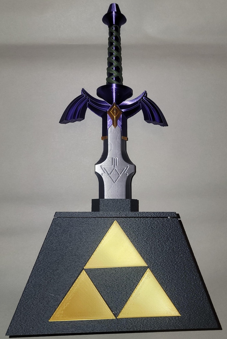 May include: A purple and silver Master Sword replica with a green rope handle, standing on a black base with a gold Triforce symbol.