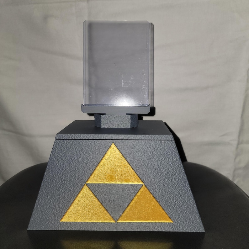 May include: A black triangular display stand with a gold Triforce symbol. A clear acrylic display case sits on top of the stand.