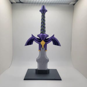May include: A purple and gold Master Sword replica with a green rope handle, displayed on a black base.
