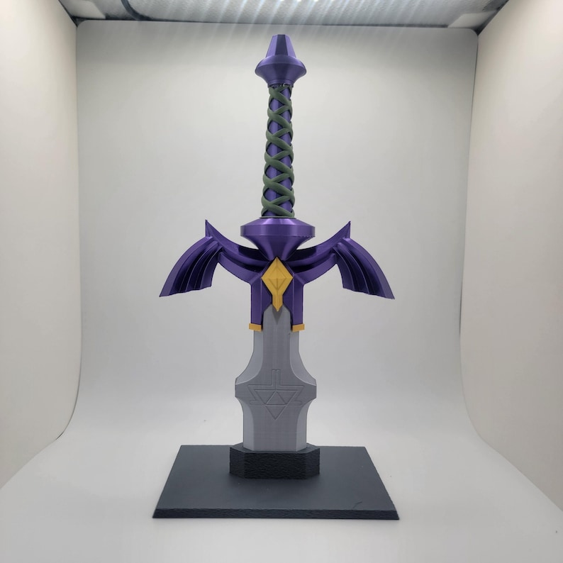 May include: A purple and gold Master Sword replica with a green rope handle, displayed on a black base.