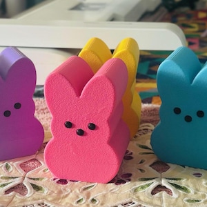 May include: A collection of colorful, bunny-shaped marshmallow treats. The bunnies are in shades of purple, pink, yellow, and blue, each with three black dot eyes. These Easter-themed candies are arranged on a patterned surface.