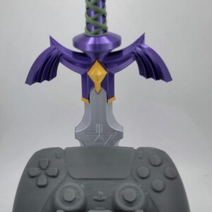 May include: A gray video game controller stand with a purple and gold Master Sword design. The stand features a triangular base with a gold Triforce symbol.