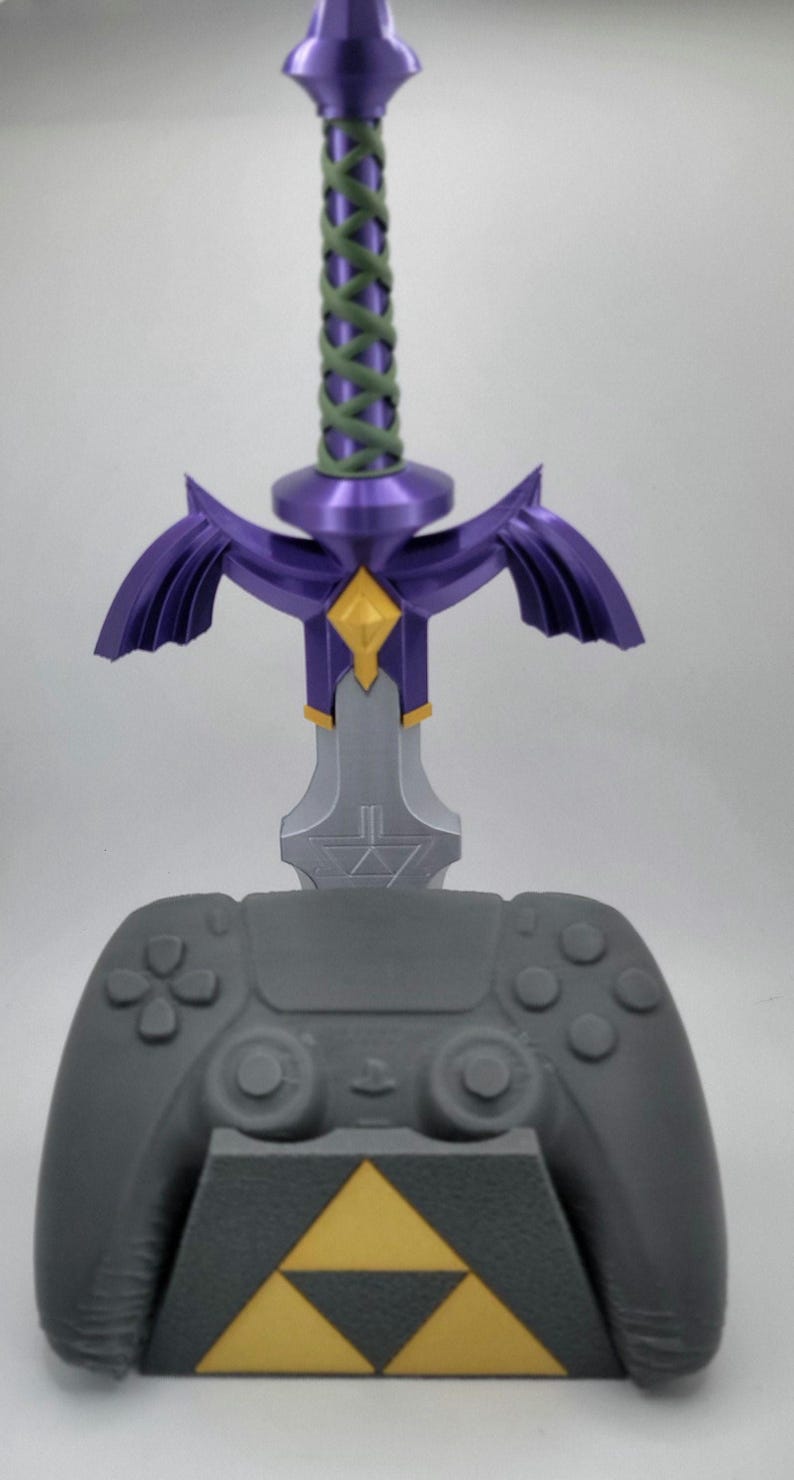May include: A gray video game controller stand with a purple and gold Master Sword design. The stand features a triangular base with a gold Triforce symbol.