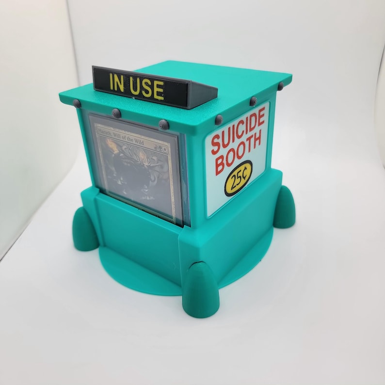Op de afbeelding: Een turquoise kaarthouder in de vorm van een cabine, met de woorden "SUICIDE BOOTH" en "25&cent;" op de zijkant. De bovenkant heeft een bord met de tekst "IN USE". Een kaart is zichtbaar in de houder.