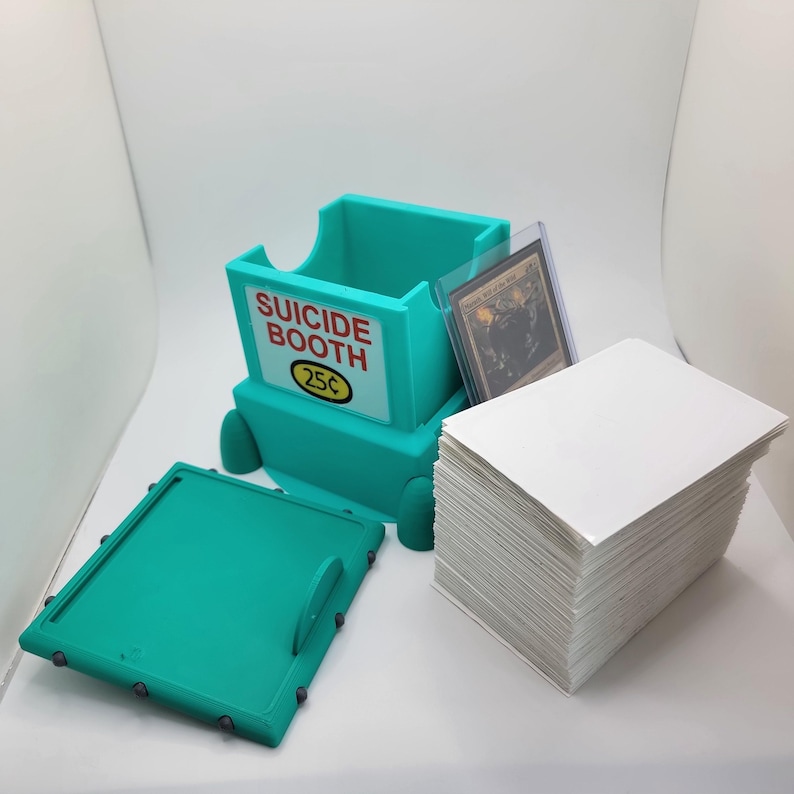 Op de afbeelding: Een turquoise kaartenopbergdoos met de woorden "SUICIDE BOOTH" en "25&cent;" op de voorkant. De doos is open, met een stapel witte kaarten en een kaart in een beschermhoesje ernaast. De deksel is open.