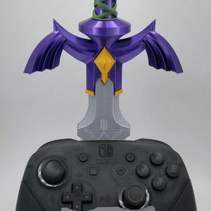 May include: A black Nintendo Switch controller is displayed on a gray stand with a gold Triforce symbol. A purple and silver Master Sword replica is mounted on the stand.