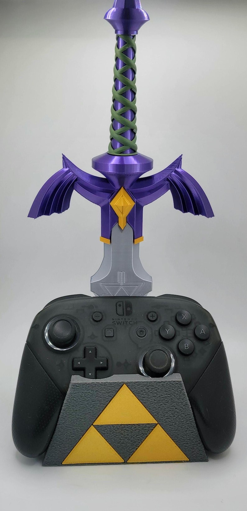 May include: A black Nintendo Switch controller is displayed on a gray stand with a gold Triforce symbol. A purple and silver Master Sword replica is mounted on the stand.