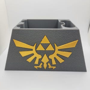 May include: A gray 3D printed game cartridge holder with a gold Triforce logo. The holder has slots for multiple cartridges.
