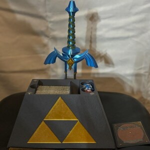 May include: A black triangular game organizer with a gold Triforce symbol. The organizer has compartments for cards and dice. A blue plastic Master Sword replica is standing upright on top of the organizer.