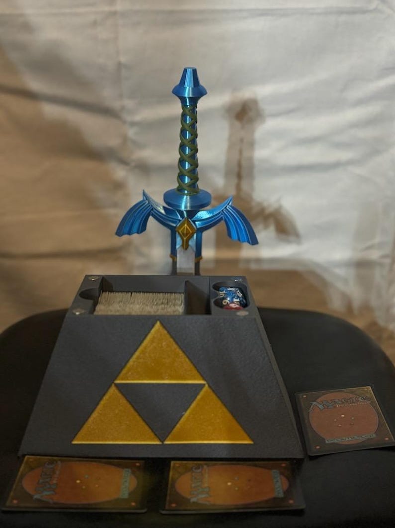 May include: A black triangular game organizer with a gold Triforce symbol. The organizer has compartments for cards and dice. A blue plastic Master Sword replica is standing upright on top of the organizer.