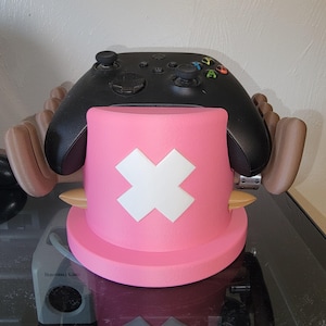 May include: A pink and white hat-shaped controller stand with a white cross design, holding a black Xbox controller. The stand has brown arms on either side. The controller has coloured buttons.