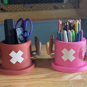 May include: Two desk organizers shaped like cartoon hats, one red and one pink, each with a white cross emblem. The red organizer holds scissors and a remote, while the pink one contains pens and pencils. The organizers have attached hand-shaped arms.