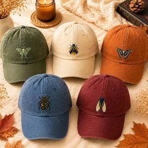 May include: Five baseball caps in olive green, beige, burnt orange, denim blue, and maroon. Each cap features a unique embroidered insect design on the front panel, including a moth, a bee, a butterfly, a beetle with moon motifs, and a cicada.
