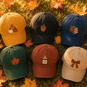 Könnte beinhalten: Sechs Baseballkappen in verschiedenen Farben wie Gelb, Anthrazit, Marineblau, Dunkelgrün, Rostrot und Beige. Jede Kappe hat ein einzigartiges gesticktes Motiv: Kürbisse, eine Eichel, ein Bücherstapel, ein Ahornblatt, eine Kerze mit dem Text COZY SEASON und eine Schleife.