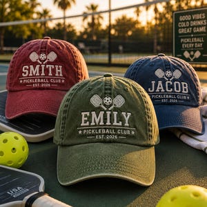 K&ouml;nnte beinhalten: Drei personalisierte Baseballkappen in Rot, Gr&uuml;n und Marineblau, jede mit einem Namen und "Pickleball Club - Est. 2026" bestickt. Die Kappen zeigen Pickleball-Schl&auml;ger- und Ballgrafiken. Ein gelber Pickleball und Schl&auml;ger befinden sich im Vordergrund.