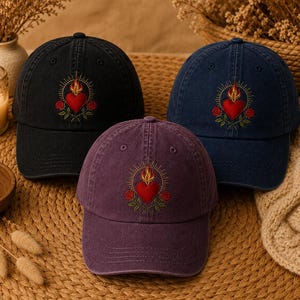 Può includere: Tre cappellini da baseball in nero, blu navy e prugna, ognuno con un ricamo di un cuore rosso con fiamme gialle e linee radianti, affiancato da due rose rosse con steli e foglie verdi centrato sul pannello frontale.