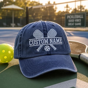 May include: A navy blue baseball cap with the text "CUSTOM NAME" and pickleball paddles and ball design. A yellow pickleball and a pickleball paddle are next to the cap. The background includes a pickleball court and a sign that says "PICKLEBALL GOOD VIBES GREAT GAME."