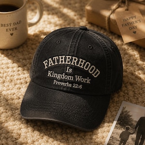 May include: A black baseball cap with the embroidered text "FATHERHOOD Is Kingdom Work Proverbs 22:6" in white. A coffee mug with "BEST DAD EVER" is visible in the background. A gift box with a "HAPPY FATHER'S DAY" tag is also in the background.
