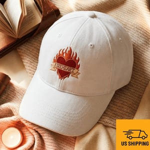 Może przedstawiać: Biała czapka bejsbolowa z czerwonym sercem i płomieniami. Na sercu znajduje się haftowany napis "FIREHEART" na bannerze. Czapka leży na beżowej powierzchni.