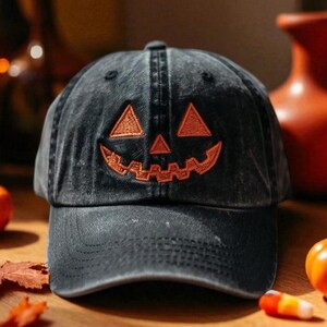 Puede incluir: Gorra de béisbol negra con una cara de calabaza naranja bordada. La gorra está sobre una superficie de madera con pequeñas calabazas, hojas y otras decoraciones de Halloween. La gorra tiene una visera curva.