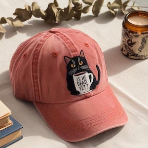 May include: A dusty rose baseball cap featuring an embroidered black cat holding a coffee mug. The mug reads "IS HE DEAD YET?". The cap has a curved brim and adjustable strap, perfect for casual wear.