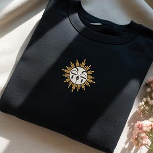 May include: Black sweatshirt with a crew neck and a sun design embroidered on the front. The sun has a smiling face with yellow rays. A cup of tea and flowers are in the background.