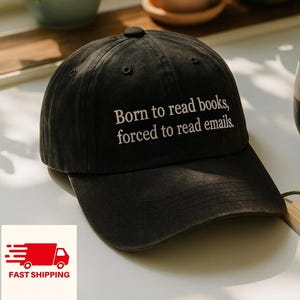 May include: Black baseball cap with the text "Born to read books, forced to read emails." The cap is sitting on a white surface next to a mug and a book.