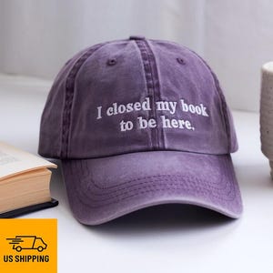 以下が含まれることがあります： 「I closed my book to be here」と刺繍された紫色の野球帽。帽子は柔らかく使い古された生地で作られており、つばがカーブしています。本とマグカップが背景にあります。