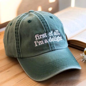 May include: A green baseball cap with the embroidered phrase "first of all, I'm a delight" in white. The hat is sitting on a wooden surface next to an open book and a small bouquet of white flowers.