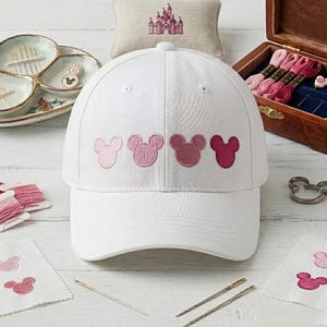 May include: A white baseball cap featuring four embroidered Mickey Mouse head silhouettes in a gradient of pink shades, ranging from light pastel pink to a deep magenta, centred on the front panel of the cap with a curved brim.