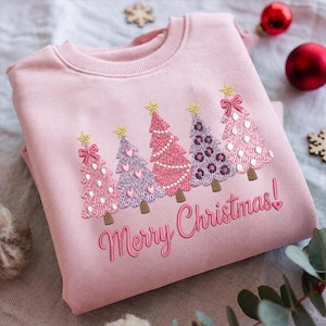 Pode incluir: Uma camisola de gola redonda rosa claro com um design bordado de cinco árvores de Natal estilizadas em tons de rosa e roxo, cada uma com uma estrela dourada no topo. O texto Merry Christmas está bordado em cursivo rosa sob as árvores.
