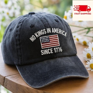 May include: A navy blue baseball cap with the text "NO KINGS IN AMERICA SINCE 1776" and an embroidered American flag. The cap is sitting on a wooden surface.