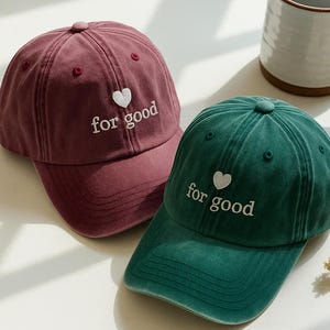 May include: Two baseball caps, one burgundy and one teal, are displayed. Each cap has a white heart and the words "for good" embroidered on the front. The caps are made of a soft material and have a classic, curved brim.
