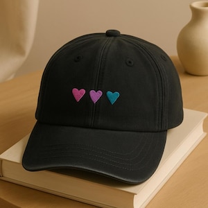 May include: Black baseball cap featuring three embroidered hearts in pink, purple, and teal. The cap rests on a closed book on a wooden table. A small, cream-coloured vase is in the background.
