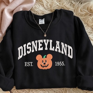 Może przedstawiać: Czarna bluza z okrągłym dekoltem z napisem "DISNEYLAND" w białych, łukowatych literach. Poniżej tekstu znajduje się pomarańczowa dynia z uszami Myszki Miki i napisem "EST. 1955."