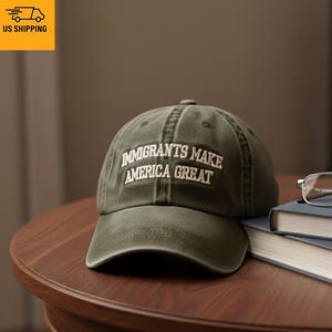 Könnte beinhalten: Olivgrüne Baseballkappe mit dem weißen, gestickten Text "IMMIGRANTS MAKE AMERICA GREAT". Die Kappe liegt auf einem Holztisch neben einem Stapel Bücher und einer Brille.