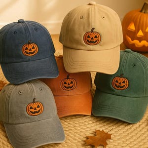 Puede incluir: Varias gorras de béisbol en tonos azul marino, beige, verde y gris, cada una con un diseño bordado de calabaza. Una calabaza tallada es visible en el fondo, lo que indica un tema de Halloween.