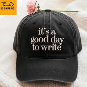 May include: Black baseball cap with white embroidered text reading "it's a good day to write". A pink flower is placed on top. The cap is made of a soft, durable material.