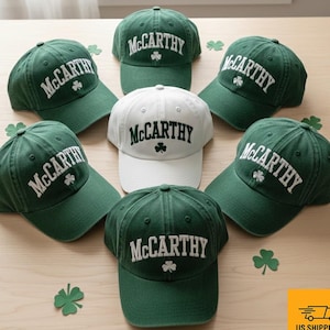 May include: A selection of green and white baseball caps, each bearing the name "McCarthy" in white lettering and a shamrock emblem. The caps are displayed on a light surface, with shamrock decorations scattered around them.